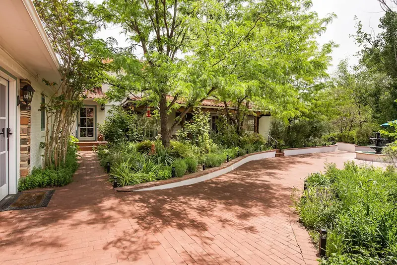 A brick patio leads to a light-colored house framed by lush trees and greenery.