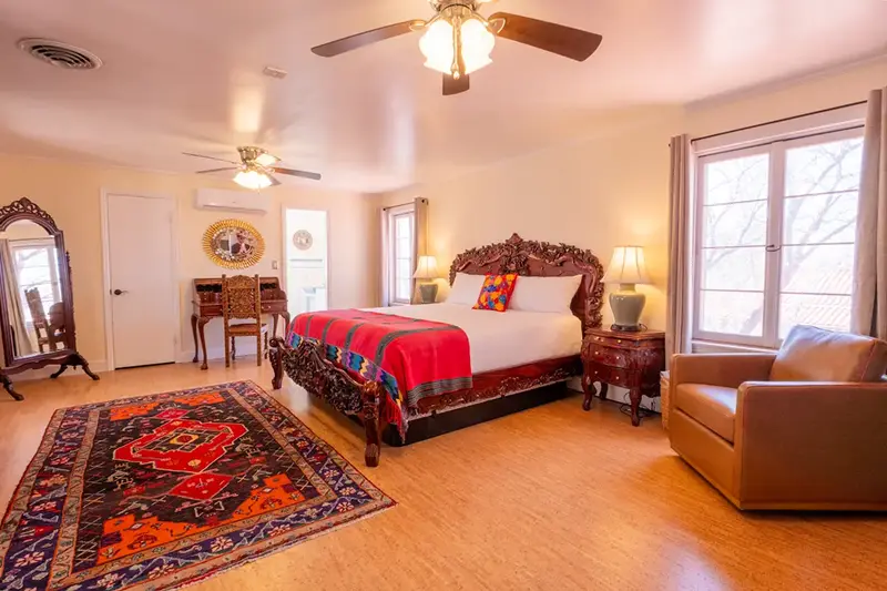 A spacious bedroom features a large, ornate bed, a patterned rug, a wooden desk, and a tan armchair.