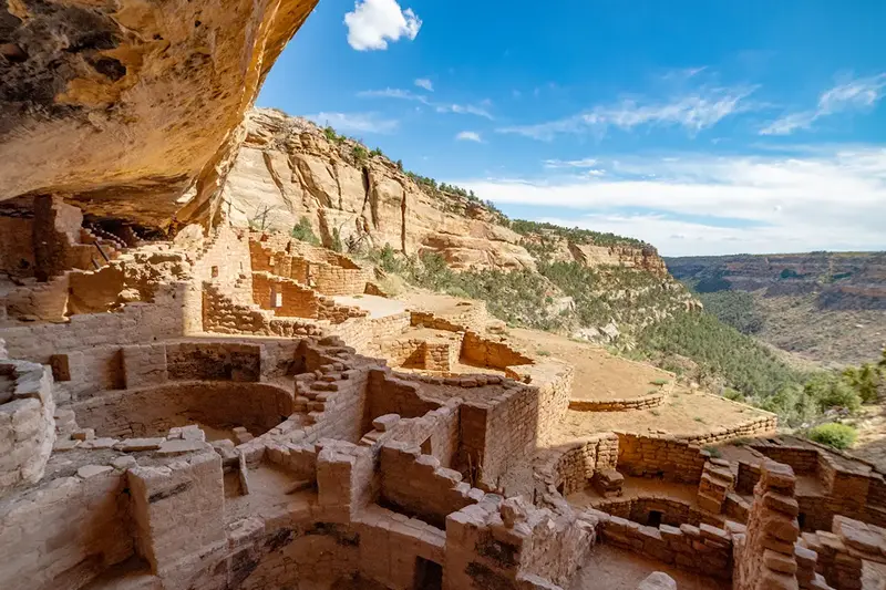 Ancient stone cliff dwellings nestled under a rock overhang, overlooking a desert canyon under a blue sky.