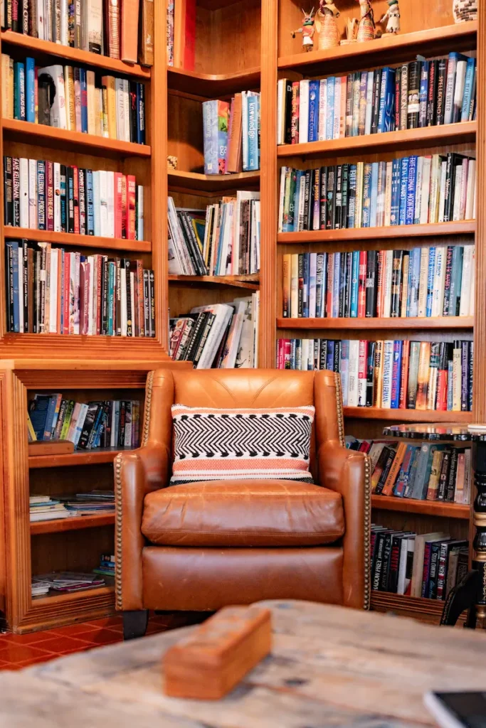 A brown leather armchair with a patterned pillow sits in front of a floor-to-ceiling wooden library bookcase.